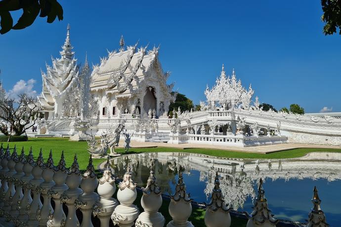Zu sehen ist der strahlend weiße Wat Rong Khun, dessen kunstvoll verzierte Fassade mit spiegelnden Glaselementen glänzt und sich majestätisch vor dem blauen Himmel erhebt.