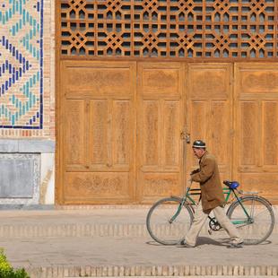 Mann schiebt Fahrrad vor großer Holztür eines Palastes, Usbekistan