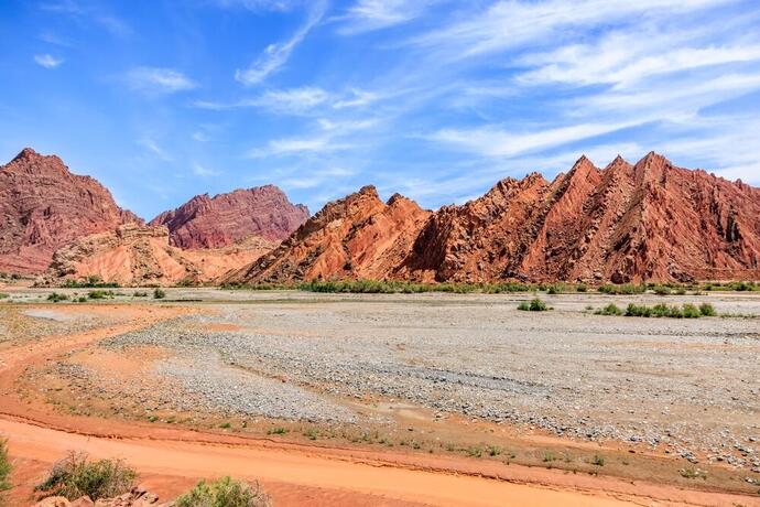 Schotterstraße führt zu einem offenen Platz vor der beeindruckenden Danxia-Berglandschaft in Xinjiang, mit roten und orangefarbenen Felsformationen.