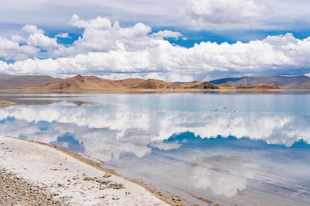 Die glatte, türkisblaue Wasseroberfläche des Yamdrok-Sees spiegelt den klaren Himmel und die umliegenden schneebedeckten Berge wider. 