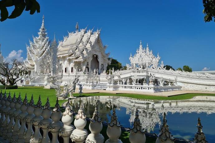 Zu sehen ist der strahlend weiße Wat Rong Khun, dessen kunstvoll verzierte Fassade mit spiegelnden Glaselementen glänzt und sich majestätisch vor dem blauen Himmel erhebt.