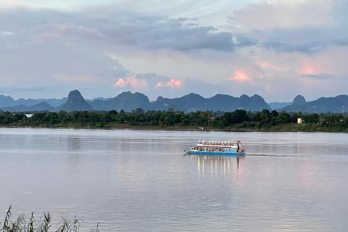 Zu sehen ist ein Schiff auf dem Mekong-Fluss bei Dämmerung. Im Hintergrund ist eine malerische Landschaft zu erkennen.